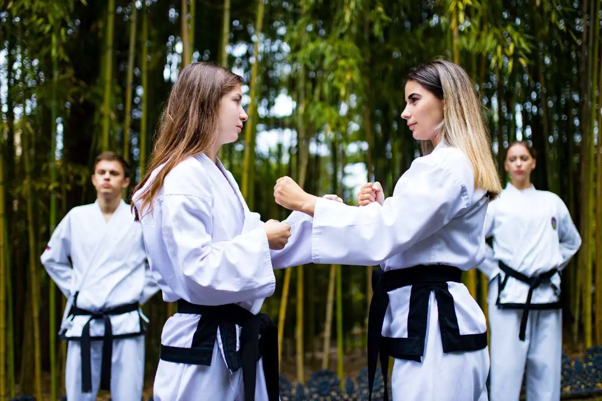Two women in white martial arts uniforms practicing hand-to-hand techniques in a bamboo forest.