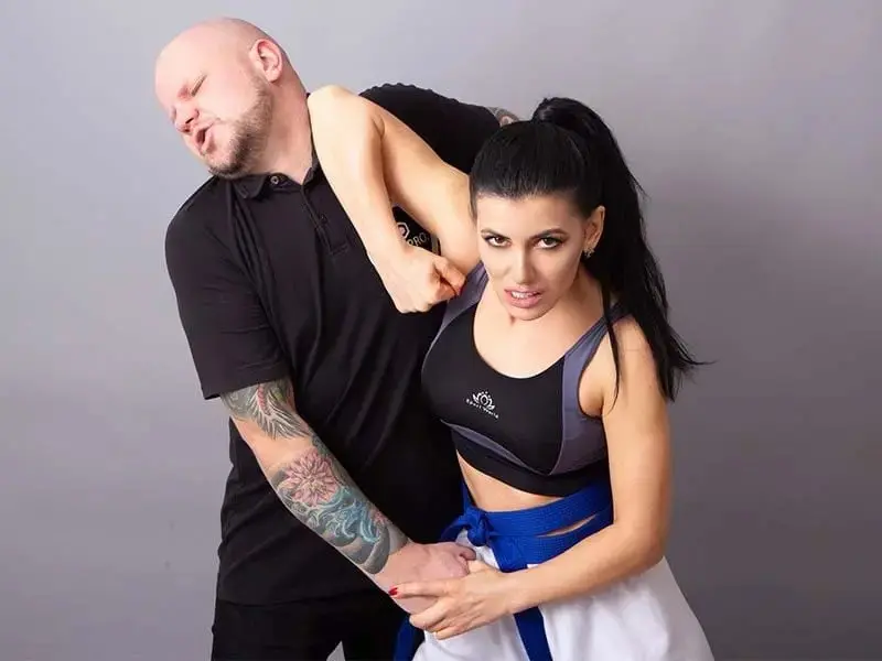 A woman in a blue belt applying a wrist-lock and arm-bar defense against a male attacker.