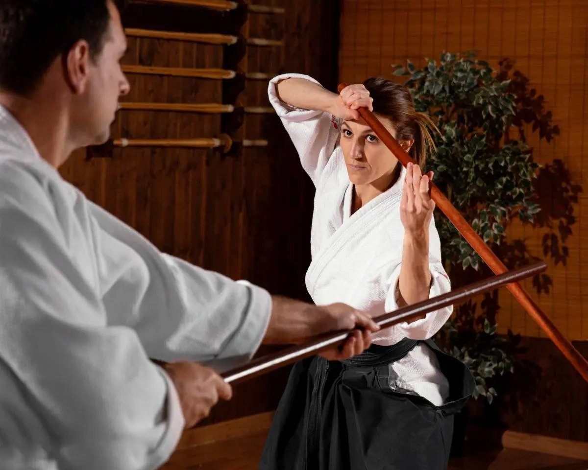 A woman in a white martial arts uniform defending against a staff attack from a male opponent.