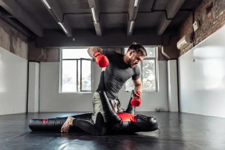 A man in red boxing gloves practicing ground strikes on a heavy grappling dummy in a gym.