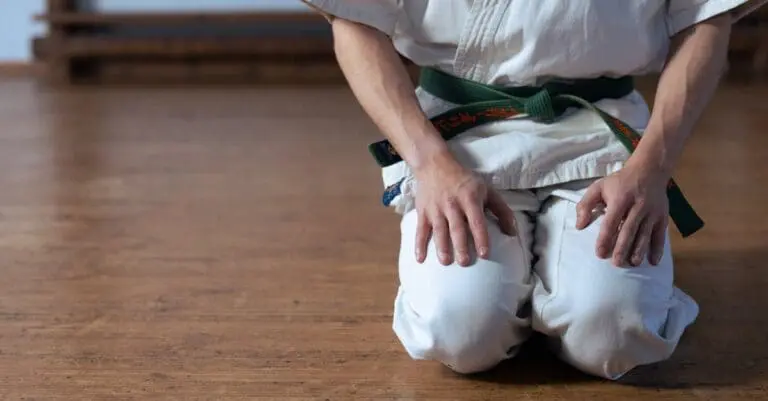 A martial artist kneeling in seiza position on a wooden floor, showcasing a green belt with custom embroidery as part of a kenpo karate belt system explained.