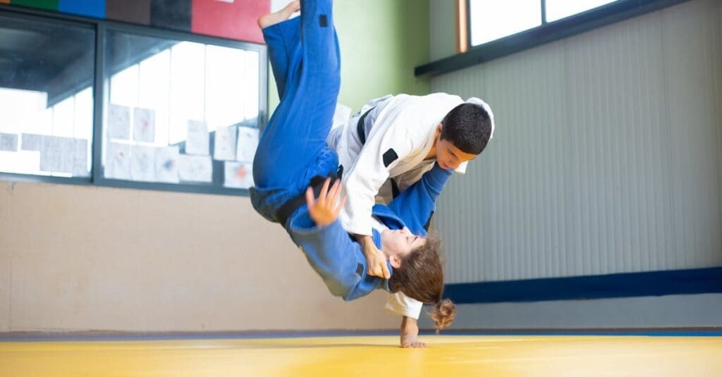 Two judo practitioners grappling on a yellow mat as one executes a ground control technique, showcasing fundamental grappling skills for a teen self defense guide.